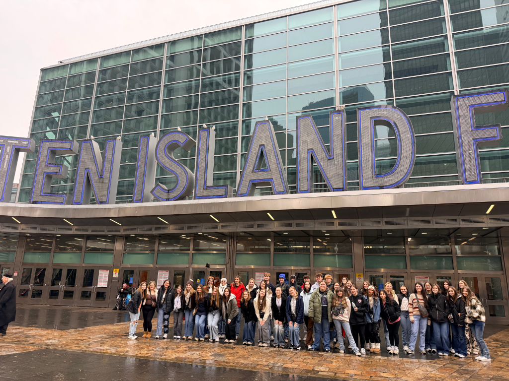 A large group of people poses for a photo in front of the Staten Island Ferry terminal, with the building’s glass facade and bold signage visible above them during their Landmark Educational Tours Registration.