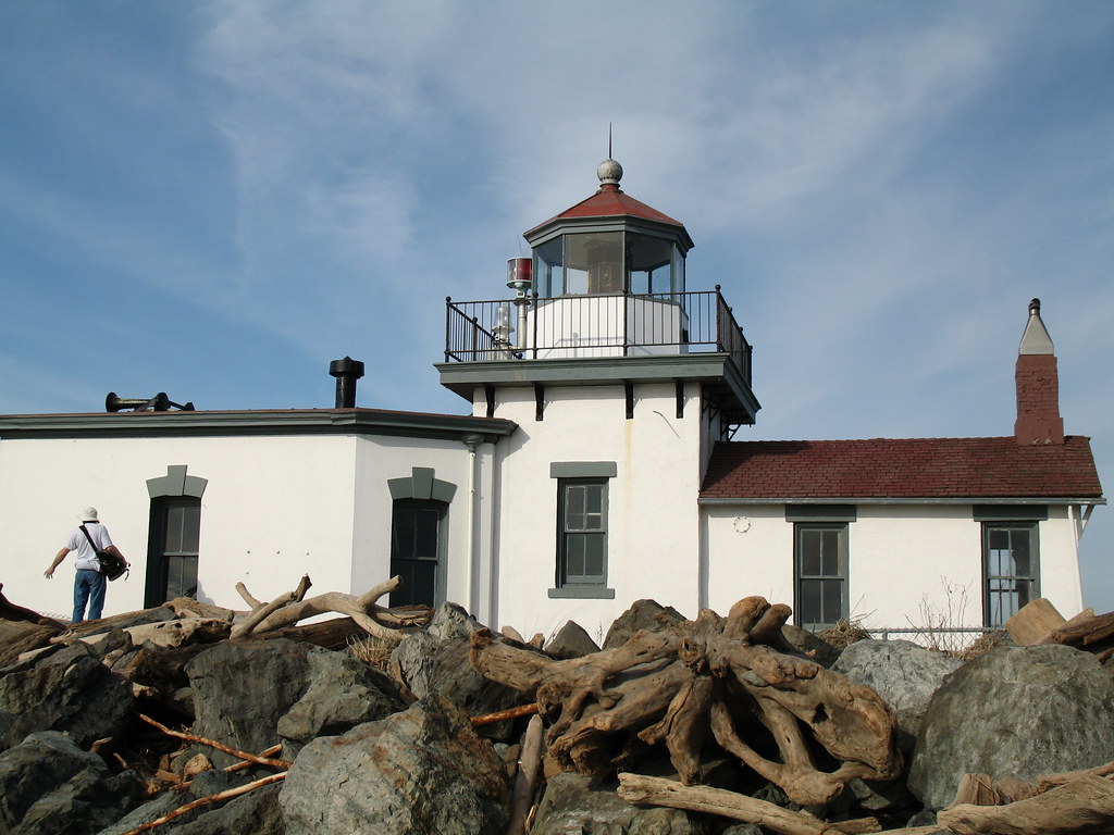 A person wearing a hat and backpack walks near a white lighthouse with a red roof, surrounded by large rocks and driftwood, under a partly cloudy sky—capturing the spirit of a Seattle School Trip adventure.