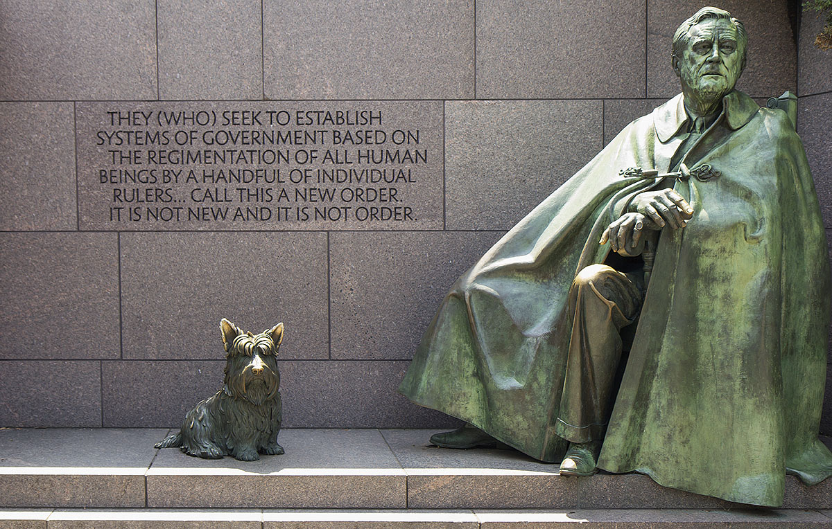 A bronze statue of Franklin D. Roosevelt seated with a cloak draped over him, next to a small dog statue, stands along the Tidal Basin Memorials Tour, set against a granite wall engraved with a quote about government and order.