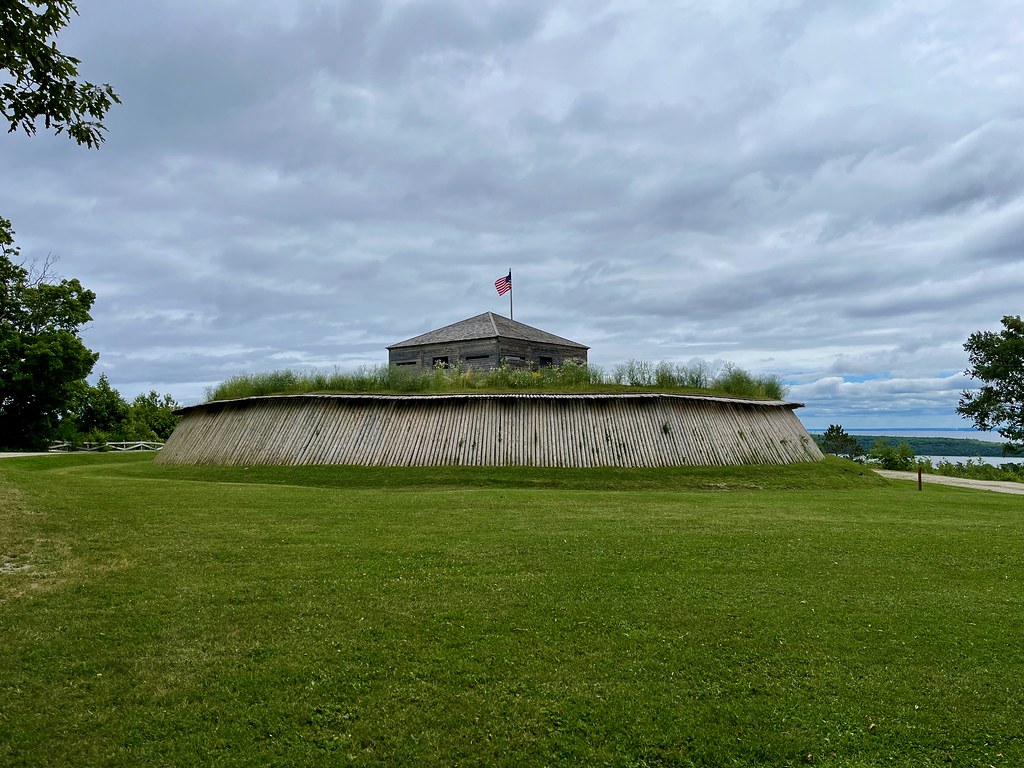 A historic stone fort with an American flag flying on top sits behind a wooden palisade, surrounded by grass under a cloudy sky—an iconic sight often visited during a Mackinac Island School Trip. Trees and water are visible in the background.