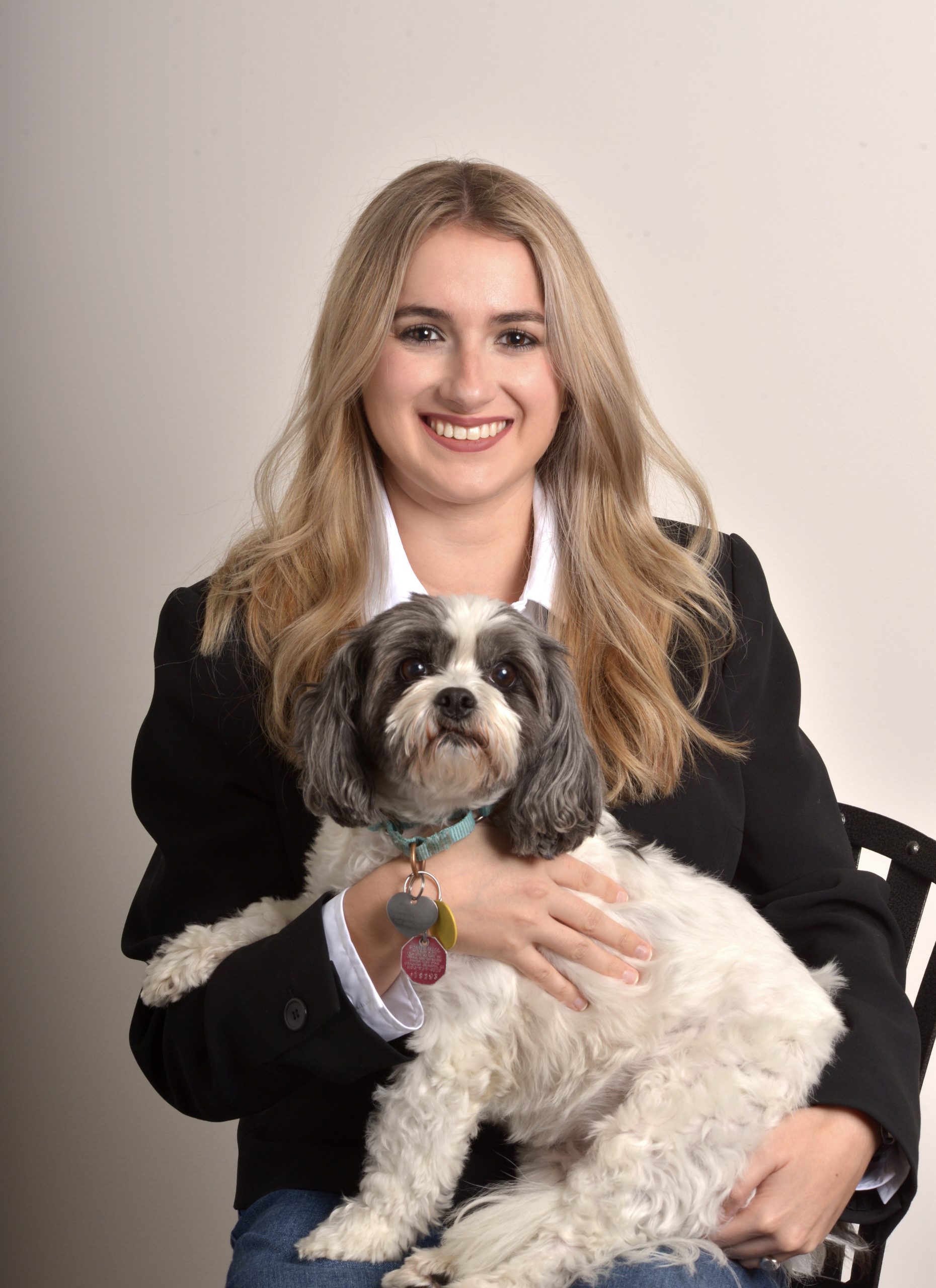 A smiling woman with long blonde hair, wearing a black blazer and white shirt, sits on a chair holding a small white and gray dog with a turquoise collar and tags in her lap. The scene is all about warmth against a plain, light-colored background.