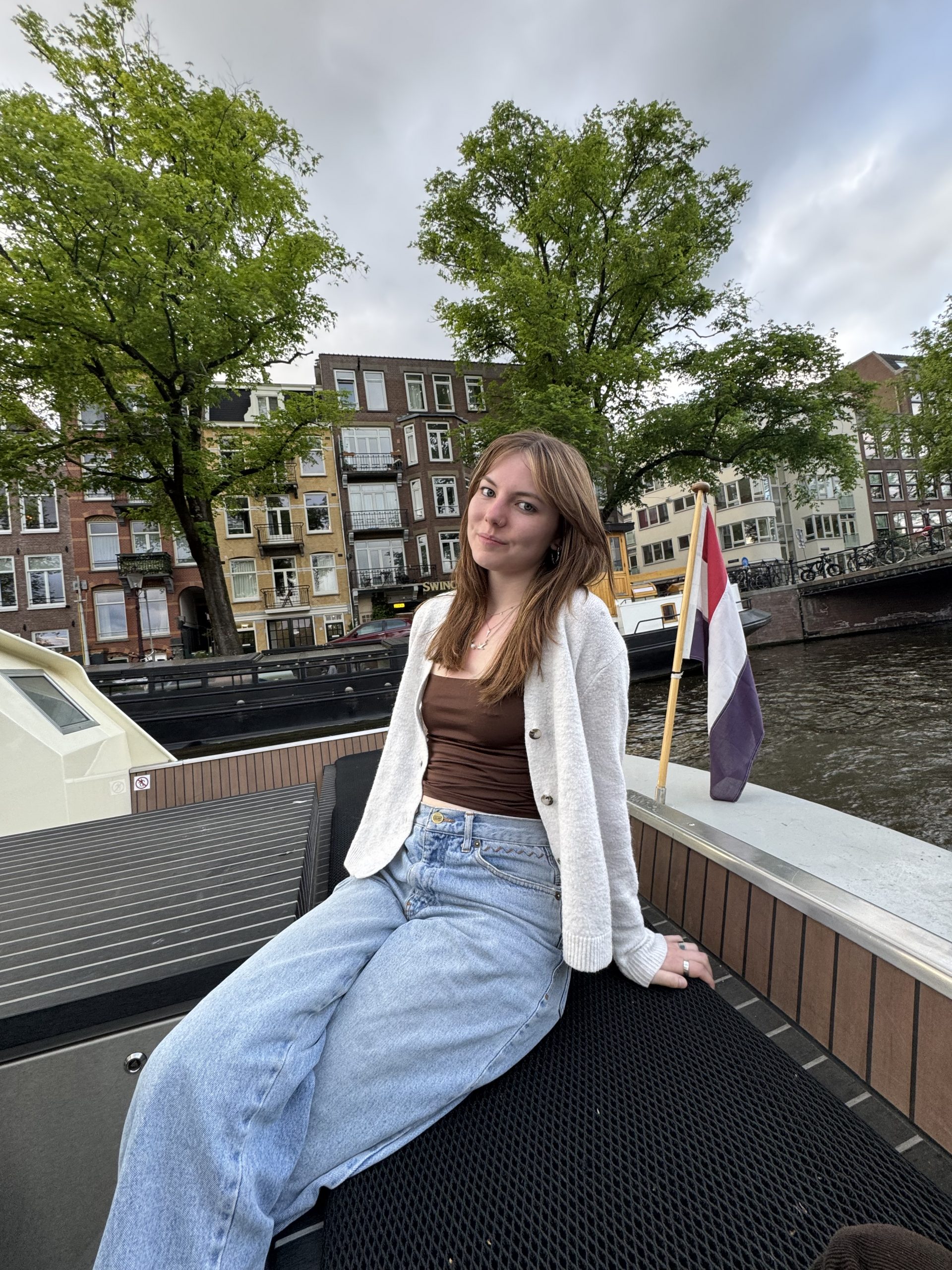 A young woman with light brown hair sits on a boat by a canal, wearing a cream cardigan, brown top, and blue jeans. Behind her are trees, canal houses, and a Dutch flag under a cloudy sky, capturing the scene's charm all about the Netherlands.