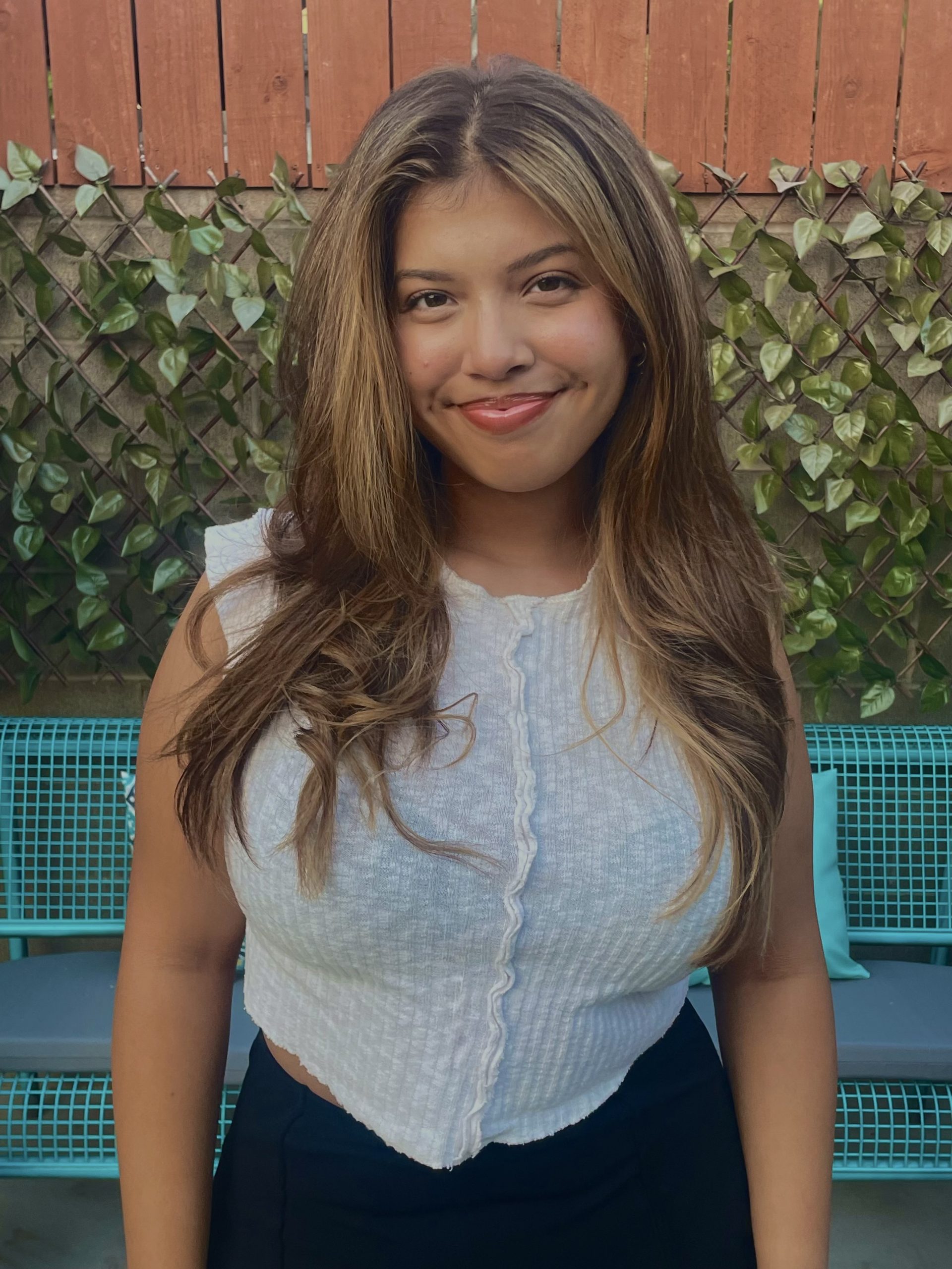 A young woman with long, wavy brown hair and a white sleeveless top smiles while standing in front of a fence with green vines and a blue bench in the background, capturing a moment about enjoying simple outdoor beauty.