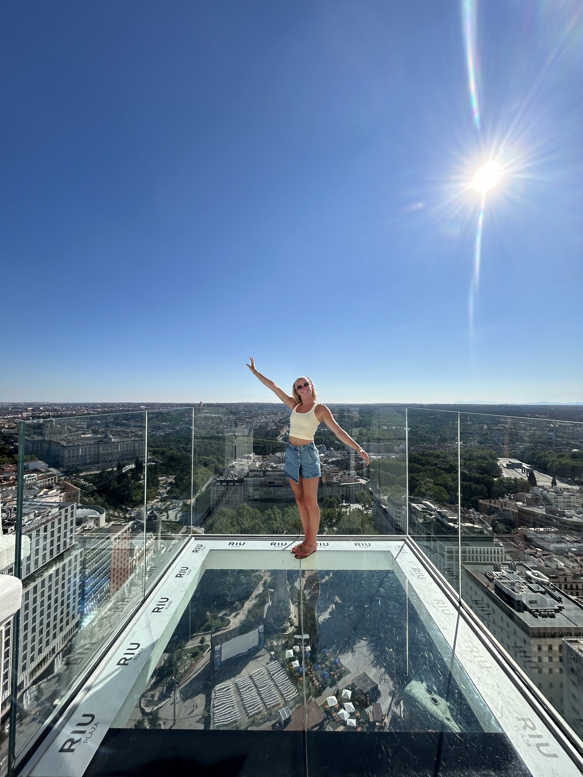 A woman stands barefoot on a glass sky deck high above the city, smiling and raising one arm. The sun shines brightly in a clear blue sky, and you can see about the city buildings and greenery stretching out below.