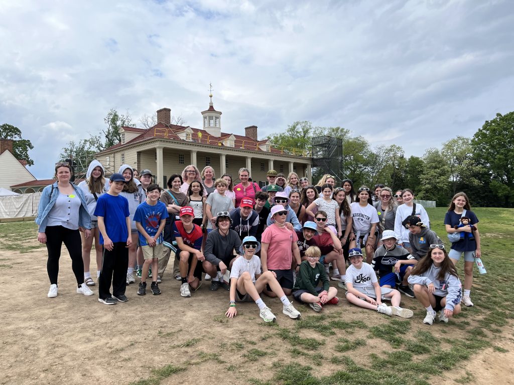A large group of children and a few adults pose for a photo outside George Washington's Mount Vernon, a historic colonial building with columns and a cupola, surrounded by trees under a cloudy sky.