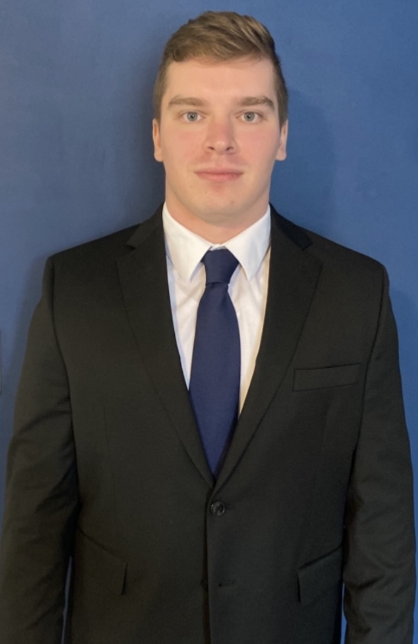 A young man in a black suit, white dress shirt, and navy blue tie stands in front of a plain blue background, looking directly at the camera with a neutral expression, as if about to share something important.