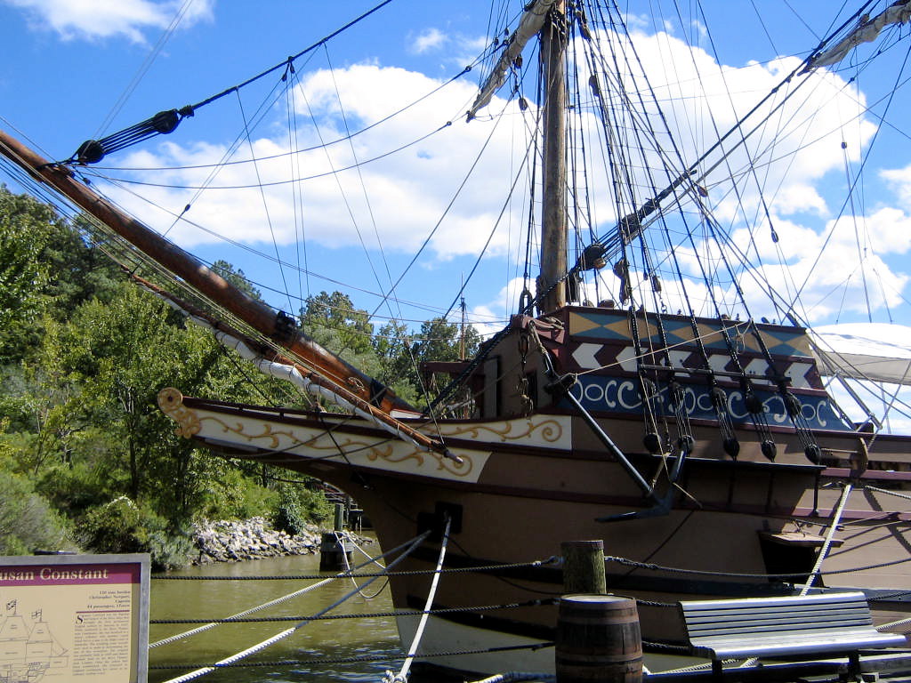 A historic wooden sailing ship with intricate designs on its bow is docked by the water, surrounded by green trees and a partly cloudy sky—an ideal stop for any Williamsburg School Trip. A sign with information stands in the foreground.