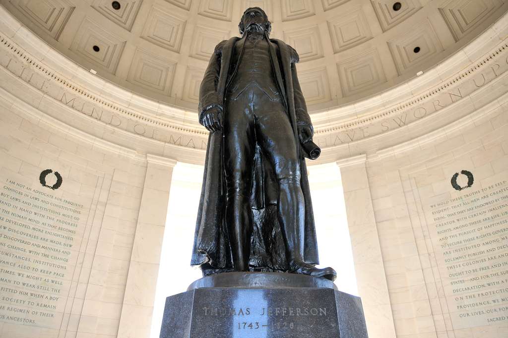 Bronze statue of Thomas Jefferson stands inside the Jefferson Memorial, part of the Tidal Basin Memorials Tour, with marble walls engraved with text and a domed ceiling above. Jefferson holds a rolled document in his hand.