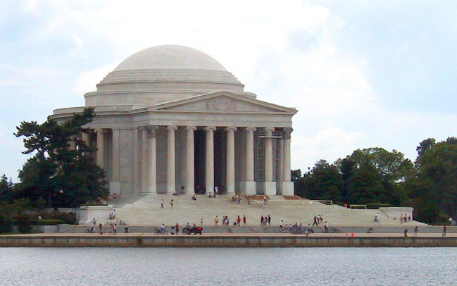 The image shows the Jefferson Memorial, a domed, columned building in Washington, D.C., with people walking on the steps and trees surrounding it, viewed from across the water—a highlight of any Tidal Basin Memorials Tour.