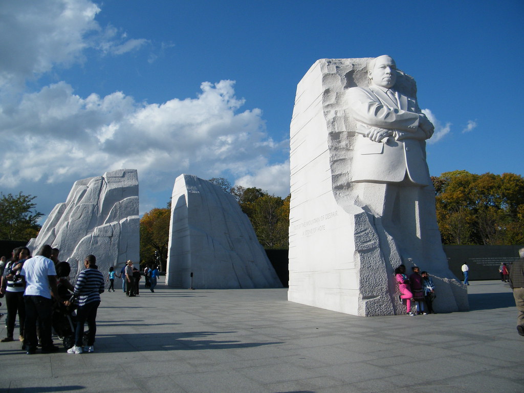 Visitors gather at the Martin Luther King Jr. Memorial in Washington, D.C., a highlight of the Tidal Basin Memorials Tour, featuring Dr. King’s stone statue standing with arms crossed against a blue sky with scattered clouds.
