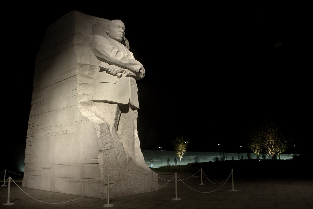 A tall stone statue of Dr. Martin Luther King Jr. stands illuminated at night with arms crossed, against a dark sky. Part of the Tidal Basin Memorials Tour, the monument is surrounded by a low barrier and trees in the background.