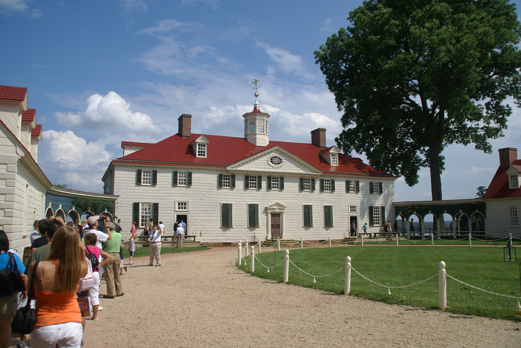 A group of people stand in line outside George Washington's Mount Vernon, a large historic white house with a red roof, green shutters, cupola, green lawn, trees, and a blue sky with scattered clouds.