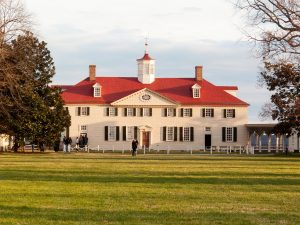 A large historic mansion, George Washington's Mount Vernon, with a red roof, white walls, and black shutters sits on a grassy lawn. Several people are walking near the entrance, and trees frame the scene.