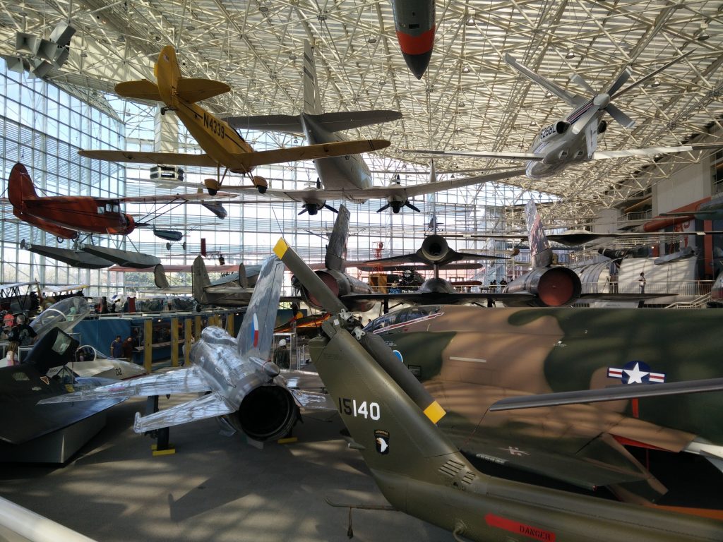 A large indoor aviation museum in Seattle showcases military aircraft suspended from the ceiling and arranged on the floor, with sunlight streaming through tall windows and Seattle Field Trip groups exploring the impressive exhibits.