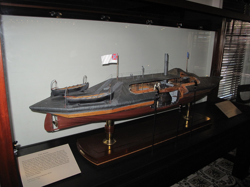 A model of an ironclad warship displayed in a glass case, featuring a Confederate flag and detailed wooden and metal components, is showcased with informational plaques—an intriguing highlight of the Savannah School Trip.