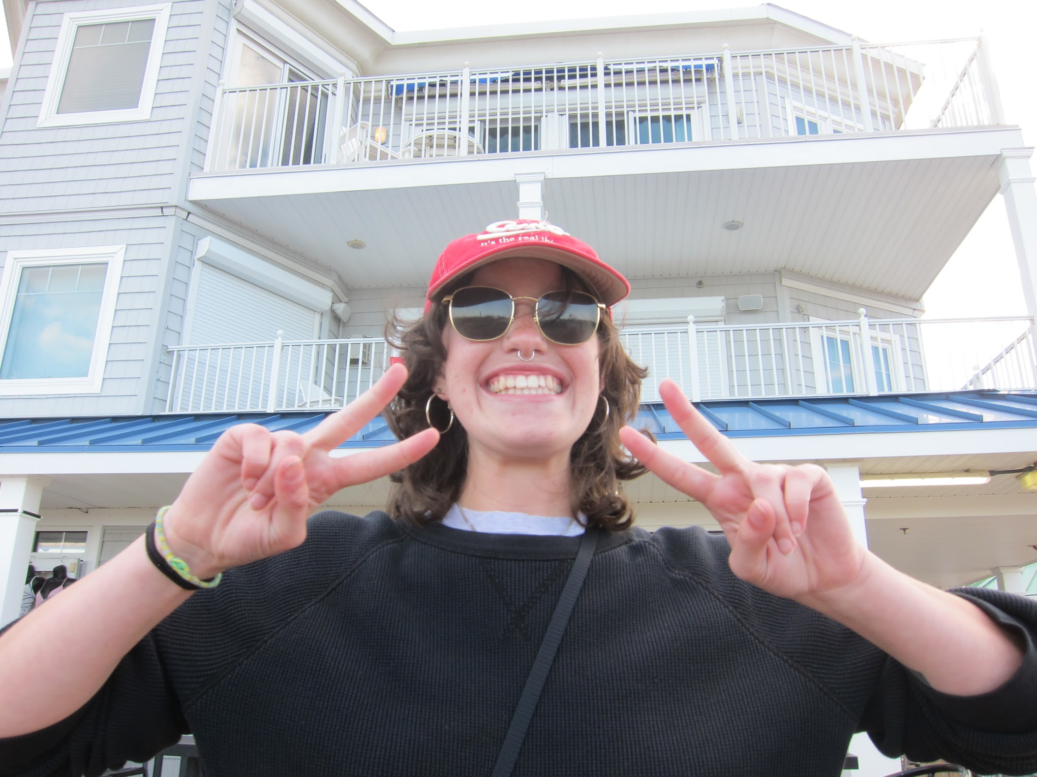 A person wearing a red cap, sunglasses, and a black sweater smiles and makes peace signs with both hands in front of a large, light gray seaside house, looking about as cheerful as can be.