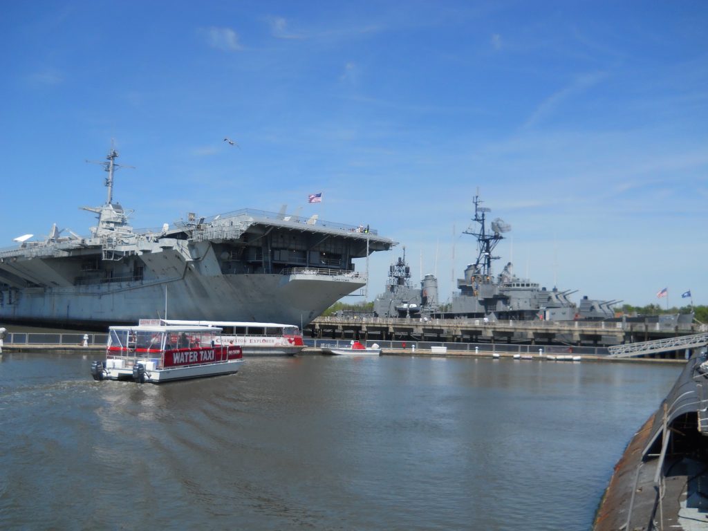 A large aircraft carrier and a battleship are docked at a naval museum, perfect for a Charleston School Trip. A red and white water taxi glides by in the foreground on a sunny day beneath a clear blue sky.