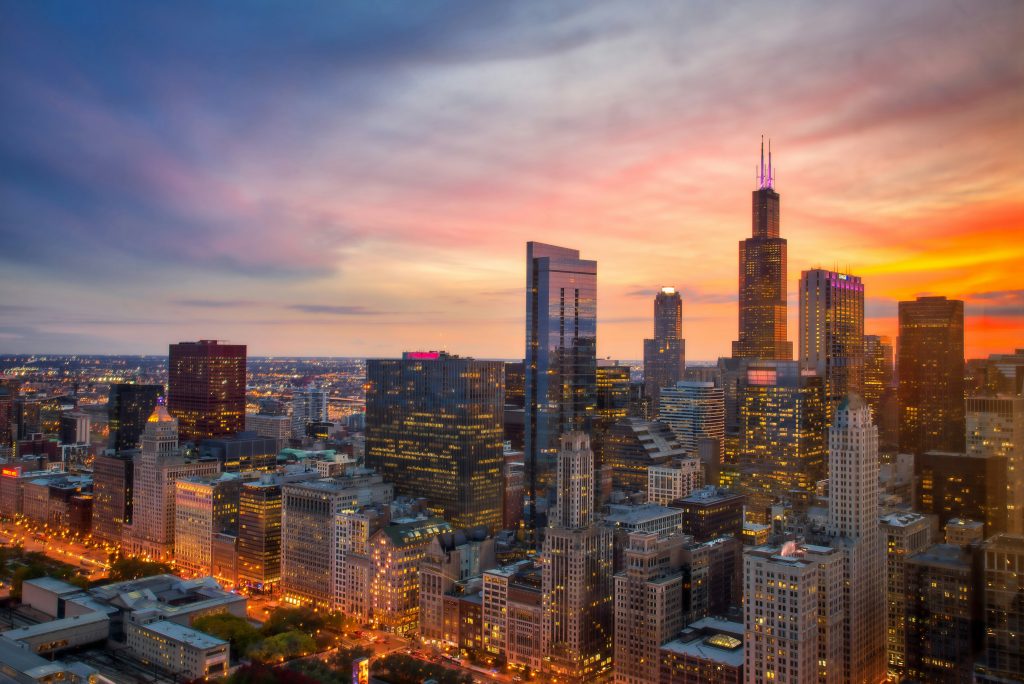 A vibrant city skyline at sunset with tall skyscrapers and colorful lights illuminating the streets below; perfect for a Chicago Student Tour, the sky displays a mix of orange, pink, and blue hues.