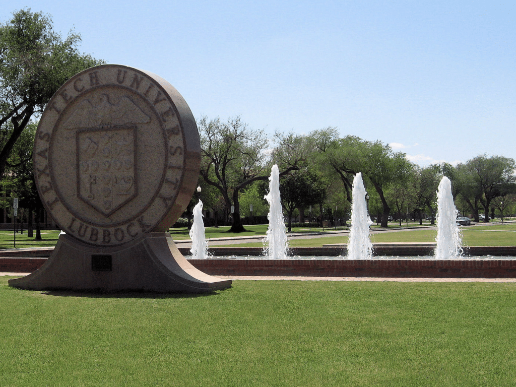 A large stone Texas Tech University seal stands as a landmark in front of a fountain with multiple jets of water on a sunny day, surrounded by green grass and trees—an inviting spot for educational tour registration.