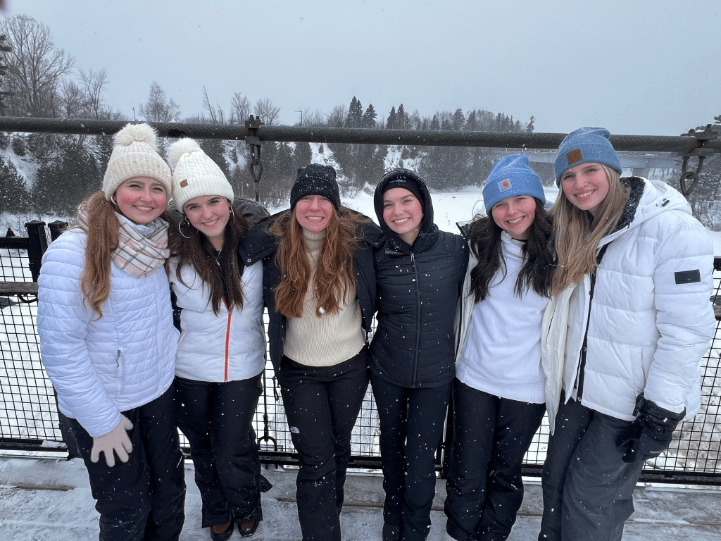 Six women dressed in winter clothing stand outside on a snowy day, smiling at the camera. Trees and snow-covered landscape are visible in the background.
