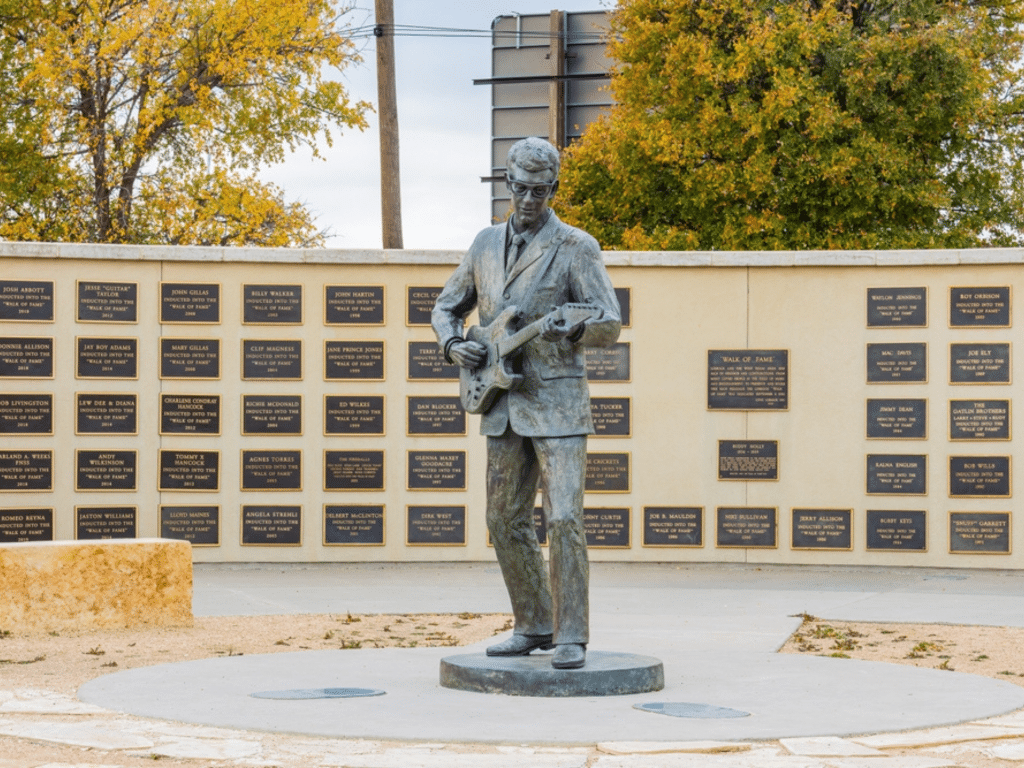 A bronze statue of a man playing an electric guitar stands in front of a curved wall covered with commemorative plaques—an iconic landmark often visited on educational tours, with trees and a road sign in the background.