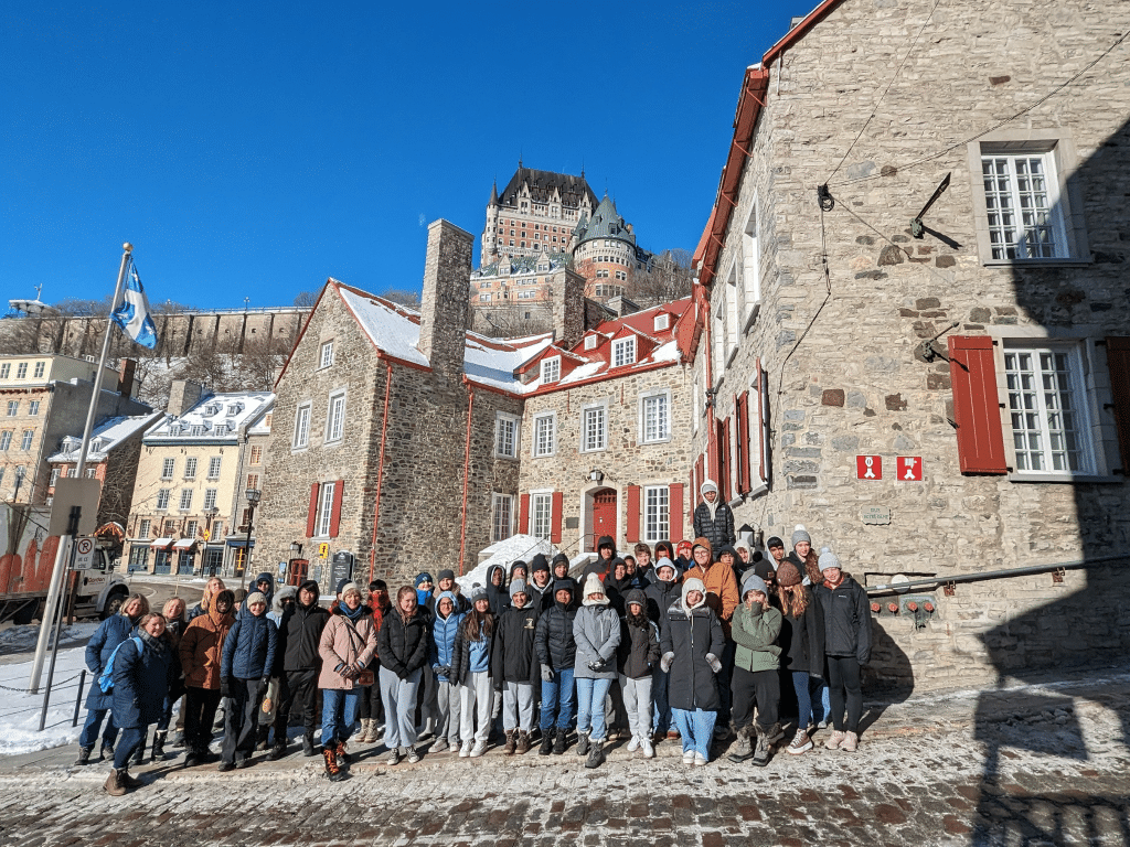 A large group of people in winter clothing poses on a cobblestone street in front of historic stone buildings with red shutters in Quebec City, with Château Frontenac visible on the hill in the background under a clear blue sky.