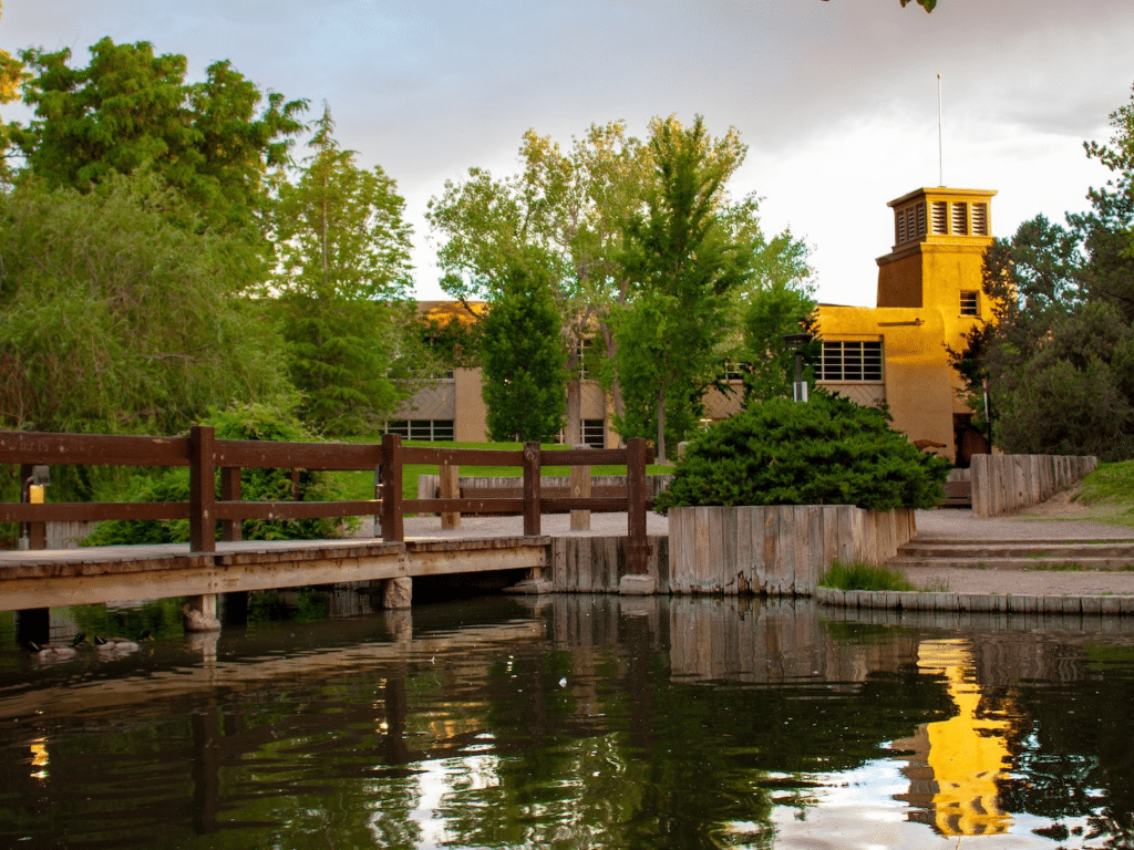 A wooden bridge crosses a pond surrounded by trees, with a tan stucco building and tower—a Landmark Educational Tour Registration site—reflected in the calm water under a cloudy sky.