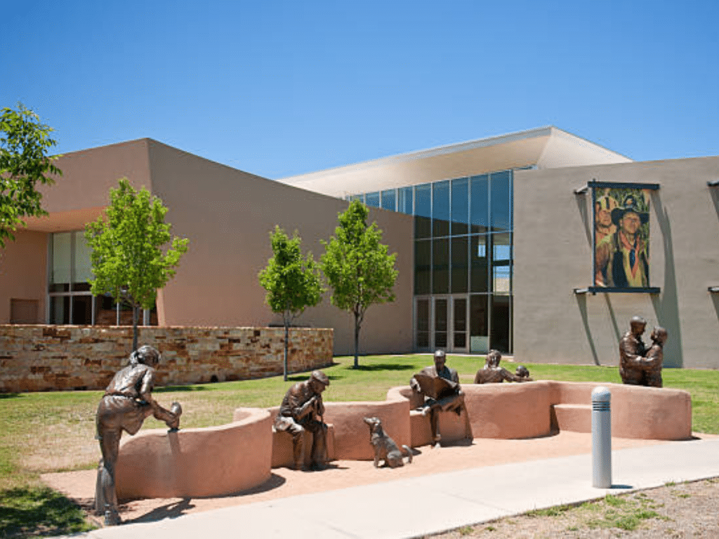 Bronze statues of people and a dog sit and stand along a curved wall outside a modern building with large windows, trees, and a mural—an inviting scene for Landmark Educational Tour Registration under the clear blue sky.