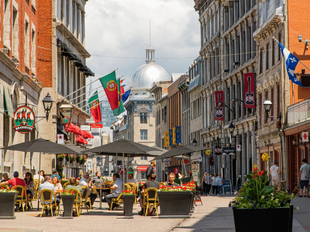 People sit at outdoor tables under umbrellas on a lively cobblestone street lined with historic buildings, colorful flags, and shops. A domed building is visible in the background under a partly cloudy sky.