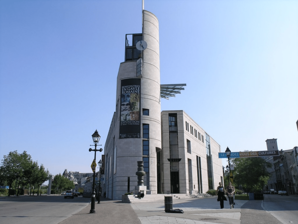 A modern, tall, cylindrical stone building with a clock and banners on its facade, located at a street corner with pedestrians and trees nearby under a clear blue sky.