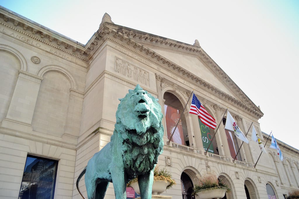 Bronze lion statue in front of the Art Institute of Chicago, with American and state flags on the classical facade under a clear sky—a must-see on any Chicago Field Trip.