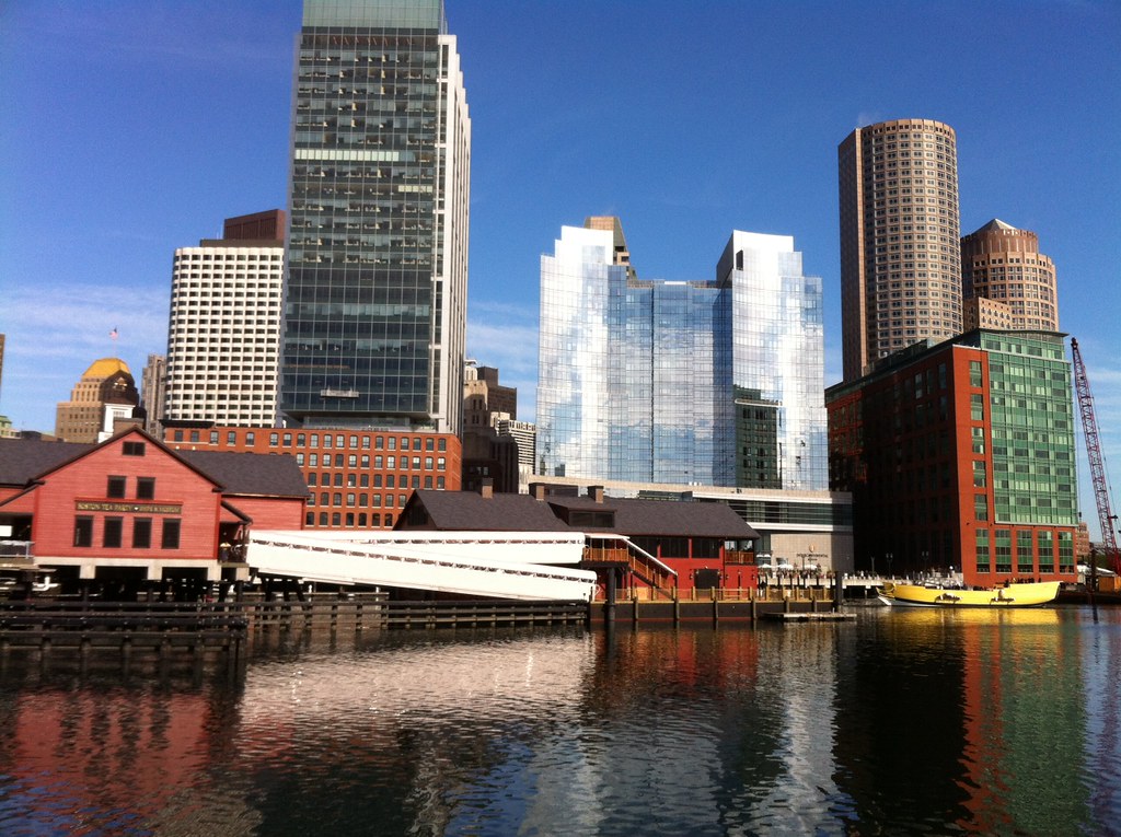 Modern high-rise buildings tower behind historic red brick and wooden structures along a waterfront, where the Boston Tea Party Ships and Museum sits reflected in the calm water. A yellow boat is docked nearby under a clear blue sky.