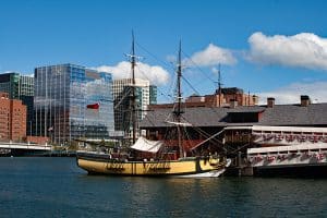 A historic wooden sailing ship, part of the Boston Tea Party Ships and Museum, is docked by a building adorned with patriotic bunting, set against modern glass offices under a blue sky with clouds.