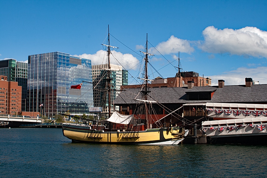 A historic wooden sailing ship, part of the Boston Tea Party Ships and Museum, is docked by a building adorned with patriotic bunting, set against modern glass offices under a blue sky with clouds.