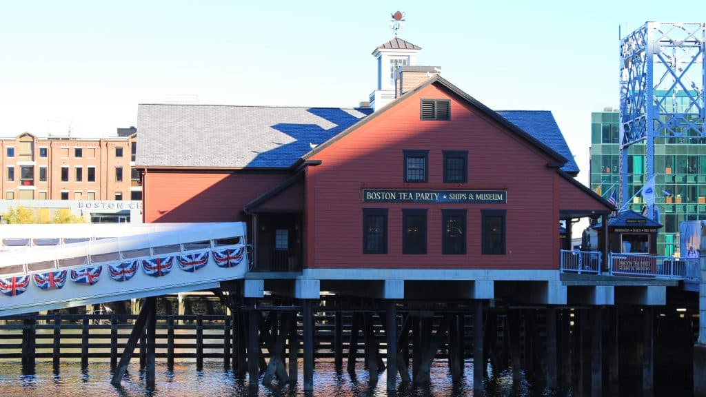 A red building labeled "Boston Tea Party Ships and Museum" sits on wooden stilts over the water, with a decorated ramp leading to it and city buildings rising in the background.