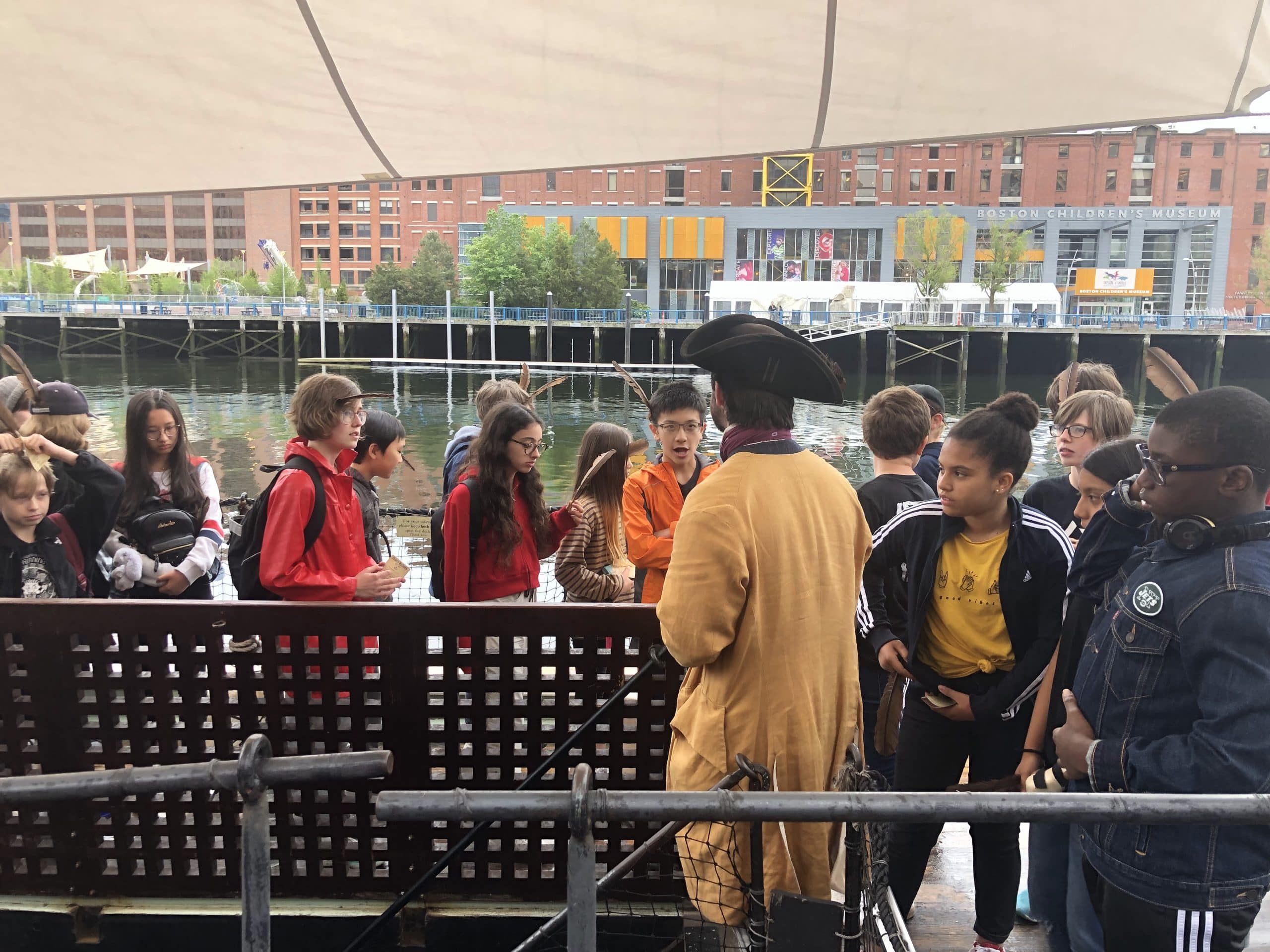 A group of children, some wearing backpacks, gather around a person in historical costume and tricorn hat on a docked ship at the Boston Tea Party Ships and Museum, with a modern brick building and water in the background.