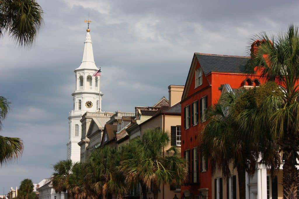 A white church steeple rises above colorful historic buildings and palm trees under a cloudy sky, capturing a classic street scene and one of the Top 10 things to do in Charleston SC.