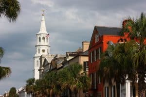 A white church steeple rises above colorful historic buildings and palm trees under a cloudy sky, capturing a classic street scene and one of the Top 10 things to do in Charleston SC.