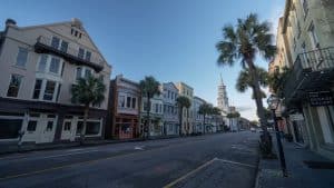 A quiet street lined with historic buildings and palm trees under a clear sky, perfect for Charleston School Field Trips, with a church steeple in the distance and a sign reading "The George Gallery" on the right.