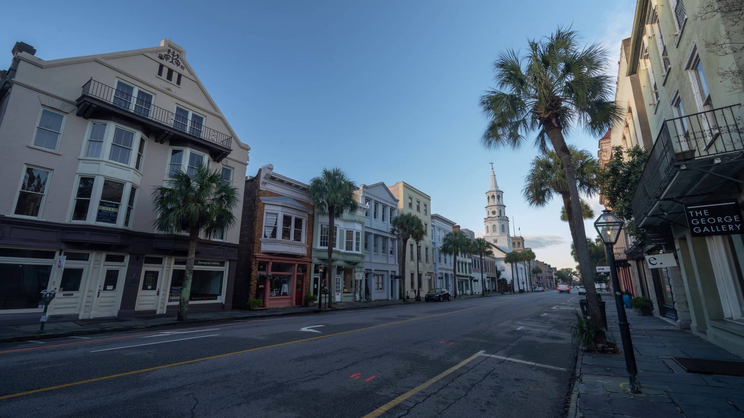 A quiet street lined with historic buildings and palm trees under a clear sky, perfect for Charleston School Field Trips, with a church steeple in the distance and a sign reading "The George Gallery" on the right.