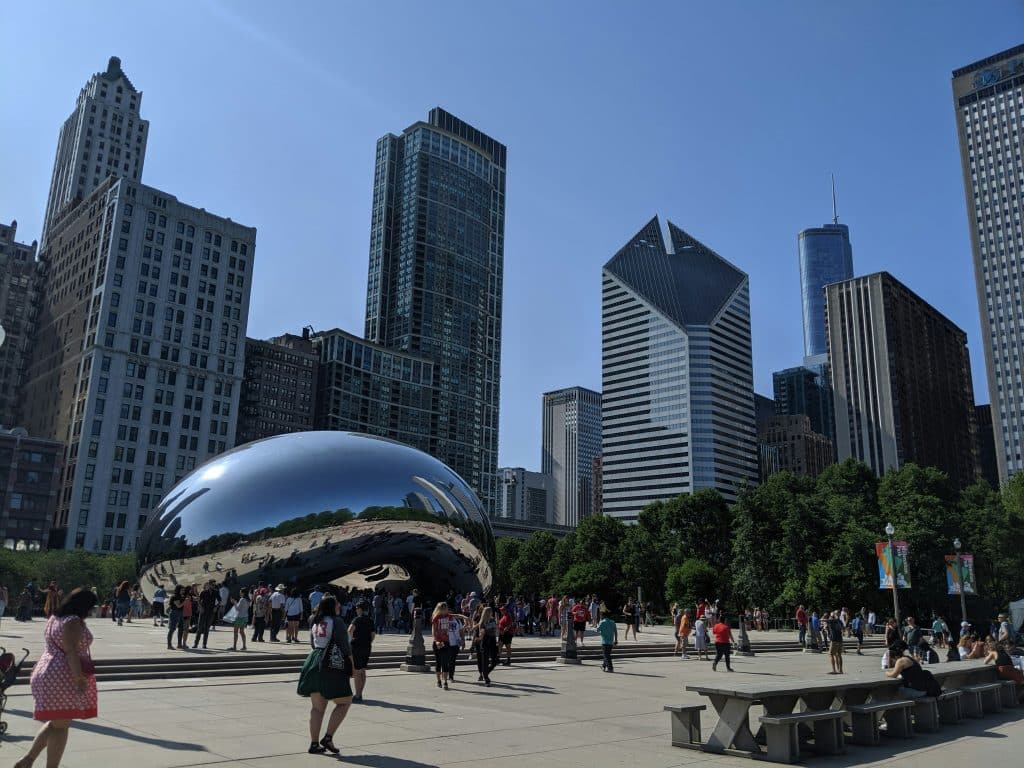 A large crowd gathers around the shiny, bean-shaped Cloud Gate sculpture in Chicago’s Millennium Park—an iconic spot for educational field trips in Chicago—surrounded by tall skyscrapers and trees under a clear blue sky.