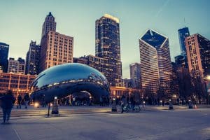 People gather around Cloud Gate, the large reflective sculpture in Millennium Park, as part of a Chicago Field Trip, surrounded by tall buildings and city lights in the evening sky.