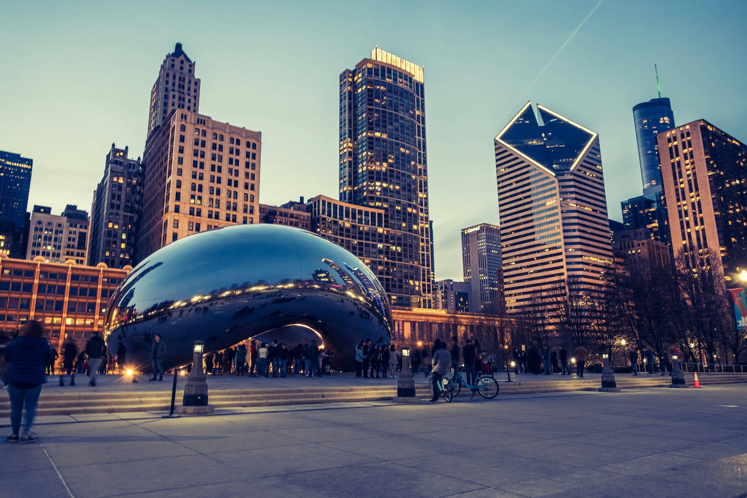 People gather around Cloud Gate, the large reflective sculpture in Millennium Park, as part of a Chicago Field Trip, surrounded by tall buildings and city lights in the evening sky.