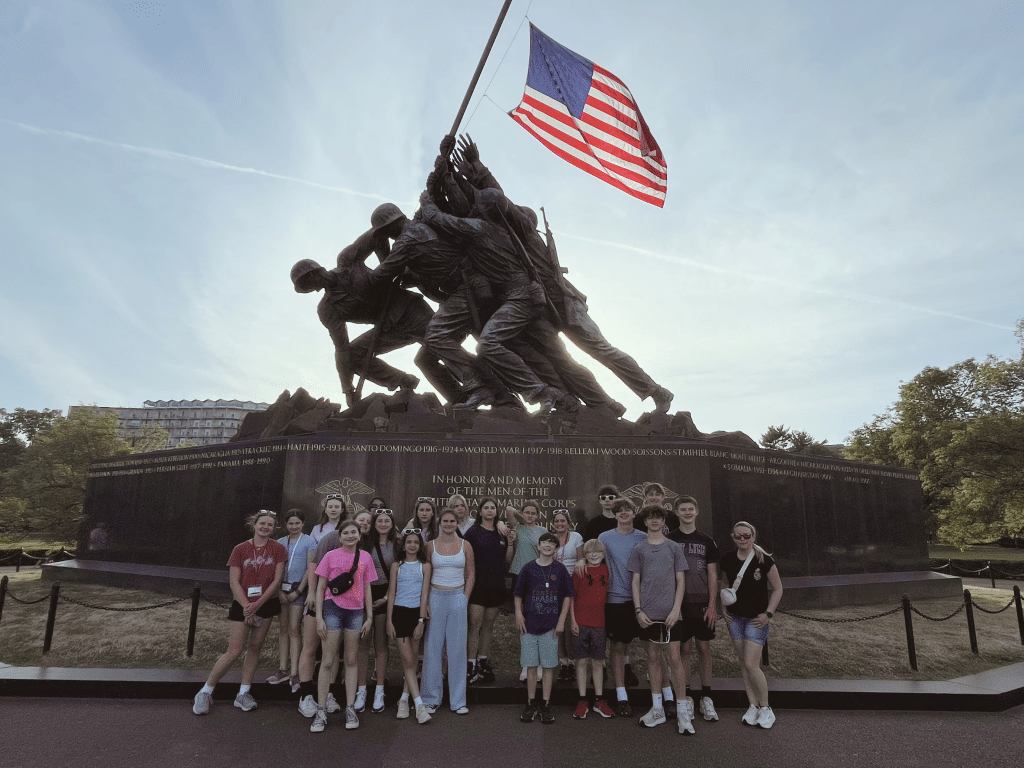 A group of people pose in front of the Marine Corps War Memorial, which depicts soldiers raising the American flag. The sky is clear and the U.S. flag waves above the statue.