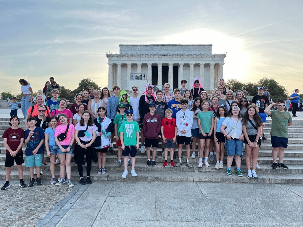 A large group of children and adults pose for a photo in front of the Lincoln Memorial in Washington, D.C., smiling on the steps with the monument visible in the background at sunset during their Landmark Educational Tours registration.