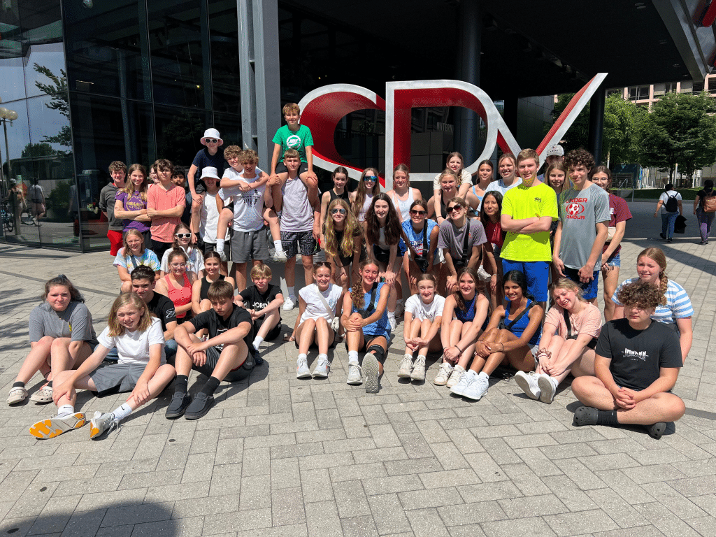 A large group of teenagers and a few adults, gathered for Landmark Educational Tours Registration, pose together outside a modern glass building with large red “CPV” letters. It is sunny, and most people are smiling and casually dressed.