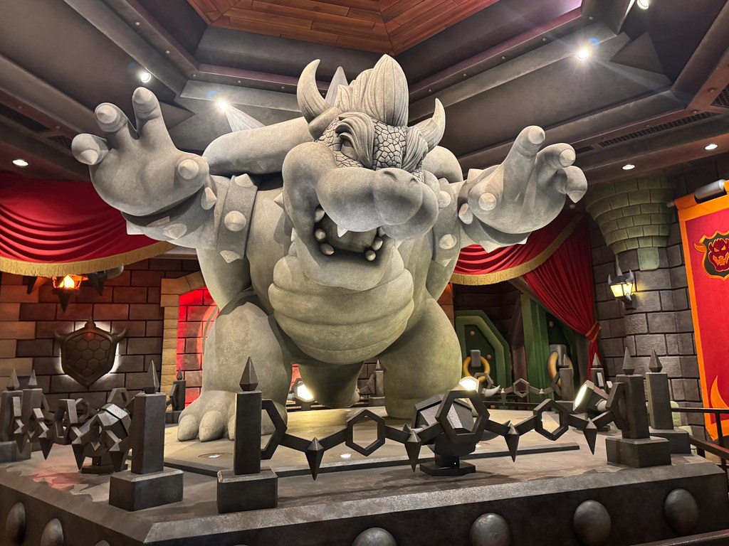 A large stone statue of Bowser, the video game character, stands with arms raised in a castle-like room—an epic sight perfect for a Los Angeles Field Trip—decorated with red curtains, stone walls, and medieval banners.