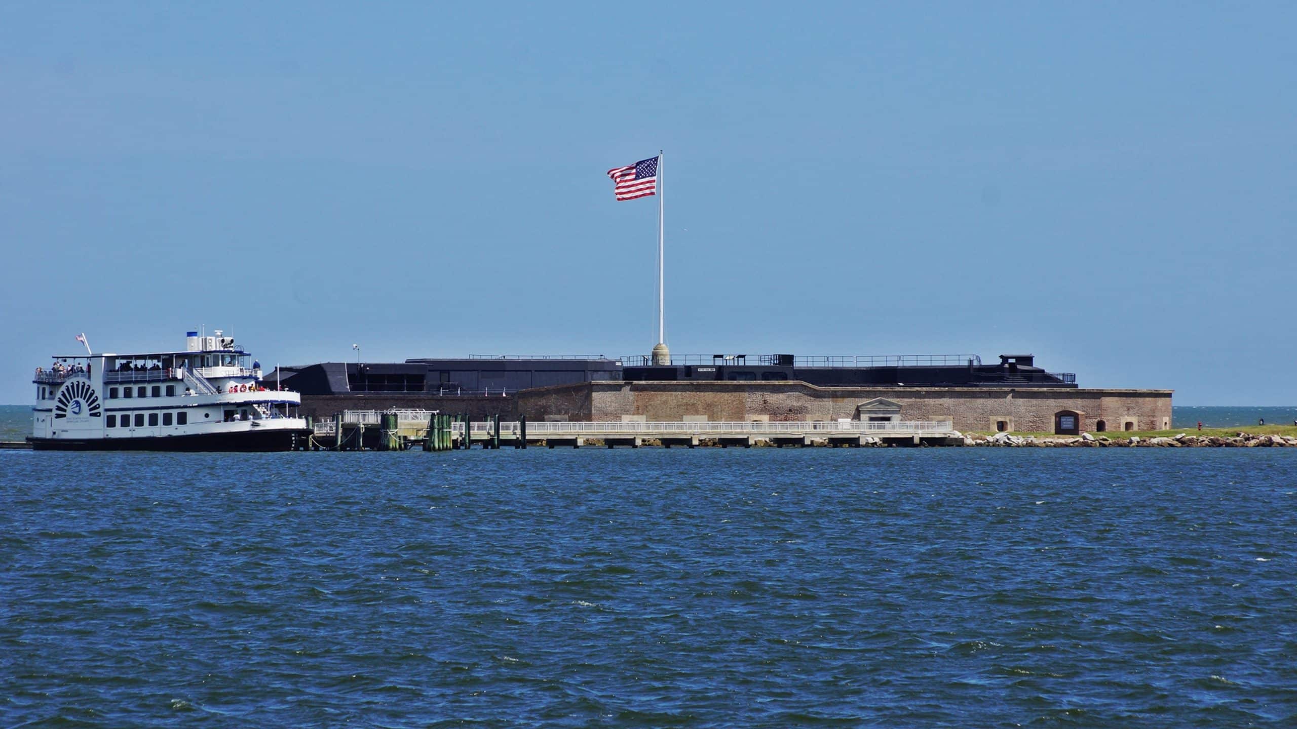 A white tour boat is docked beside a historic brick fort, one of the iconic historic landmarks in Charleston SC, with an American flag flying on a tall flagpole under a clear blue sky.