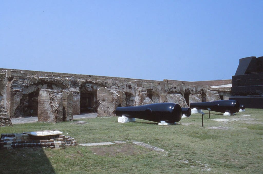 This historic brick fort, one of the notable historic landmarks in Charleston SC, features arched walls—some partially ruined—and large black cannons displayed on a grassy area beneath a clear blue sky.