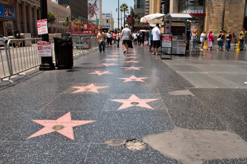People walk along the Hollywood Walk of Fame on a sunny day, passing pink stars and a damaged section of sidewalk. Buildings and barriers are visible—just one highlight you might see on the Best Educational Tours in California.